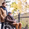 Pflegebedürftiger Mann im Rollstuhl schaut von einem Balkon in die Ferne, Pfeleger hinter ihm hält den Rollstuhl