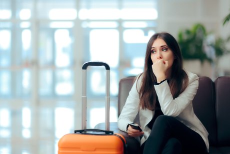 Junge Frau mit Handy sitzt im Flughafen mit Handy in der Hand, vor ihr steht ein Koffer