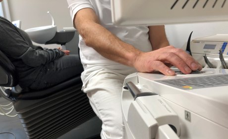 Mann in weißer Kleidung an einem Gerät mit Monitor, daneben zahnärztliche Instrumente, hinter ihm Patient im Behandlungsstuhl