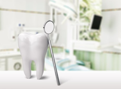 großer weißer Zahn an dem ein Mundspiegel lehnt, im Hintergrund unscharf ein Behandlungsstuhl