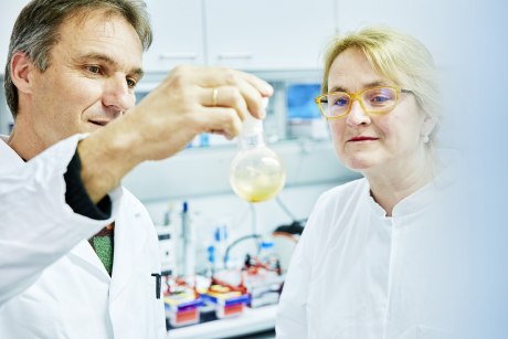 ein Mann und eine Frau in weißen Laborkitteln betrachten eine gelbliche Substanz in einem Glaskolben